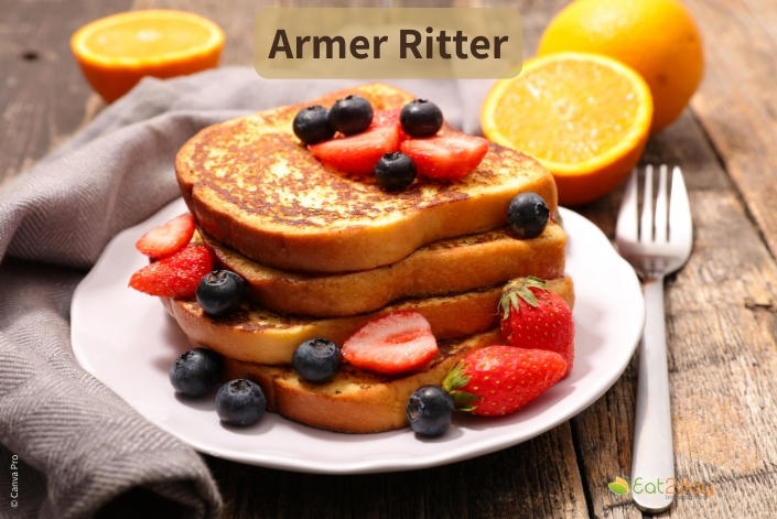 Ein Teller mit gestapelten, goldbraun gebratene Brotscheiben, belegt mit Erdbeerscheiben und Heidelbeeren. Daneben liegen halbierte Orangen und eine Gabel auf einem Holztisch. Oben im Bild steht der Titel ‚Armer Ritter‘.
