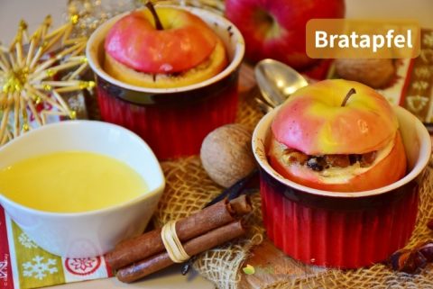 Daneben befinden sich eine Schüssel mit Vanillesauce, Zimtstangen und frische Äpfel. Oben rechts steht der Titel „Bratapfel“.