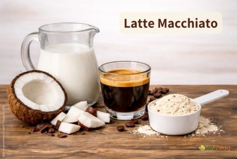 Milchkrug, Espresso und Kokosstücke neben einem Messlöffel mit Proteinpulver auf einem Holztisch. Oben rechts steht der Titel „Latte Macchiato“.