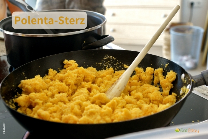 Eine Pfanne auf einem Herd mit frisch zubereitetem Polenta-Sterz, einer grob gekrümelten Maisgrießmasse, die mit einem Holzlöffel umgerührt wird. Im Hintergrund steht ein Kochtopf. Oben im Bild steht der Titel ‚Polenta-Sterz‘.
