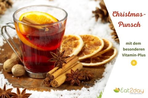 Ein Glas mit rotem Christmas-Punsch, garniert mit einer Orangenscheibe und einer Zimtstange. Daneben liegen getrocknete Orangenscheiben, Sternanis, Zimtstangen und Muskatnüsse auf einer winterlich dekorierten Oberfläche mit etwas Schnee. Rechts im Bild steht der Text ‚Christmas-Punsch mit dem besonderen Vitamin-Plus‘.