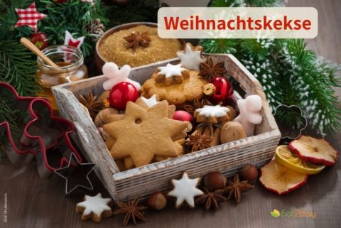 Holzkiste mit verschiedenen Weihnachtskeksen in Sternform, dekoriert mit Nüssen, Gewürzen und roten Weihnachtskugeln; darüber steht „Weihnachtskekse“.