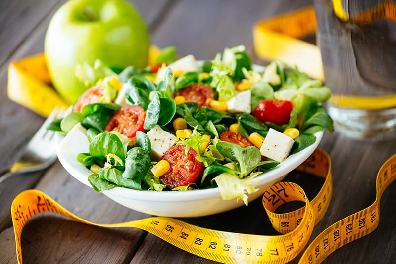 Salatteller mit Vogerlsalat, Mais, Tomaten und Schafkäse. Daneben ist ein gelbes Maßband, ein grüner Apfel und ein Gals Wasser.